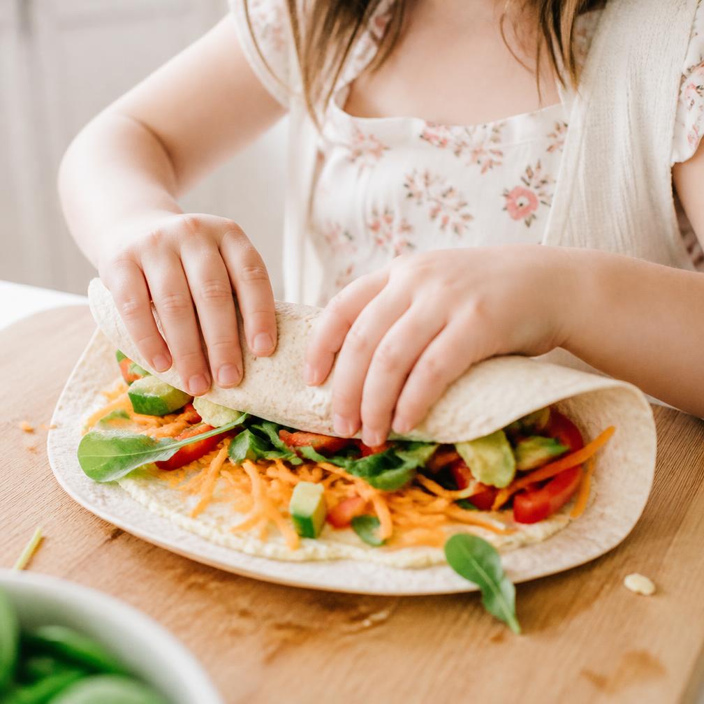 Close-up of a child's hands rolling a vegetable-filled wrap on a wooden board
